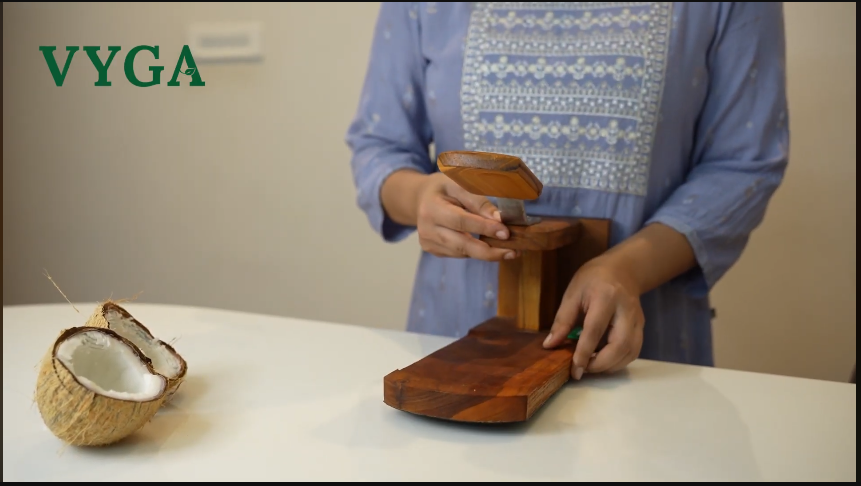 Load video: The video shows a person using the Vyga Coconut Scraper, a handcrafted wooden kitchen tool made from teak wood. The person places half a coconut on the scraper’s sharp iron blade and smoothly scrapes the white coconut flesh into a bowl. The base of the scraper has a black rubber sheet to prevent slipping, and a round groove holds the plate firmly. As the person scrapes, the narrator explains the product’s features — including the sturdy teak wood body, protective wooden cap for the blade, and its traditional design made in Kerala. The overall tone highlights its durability, safety, and elegant handcrafted finish.